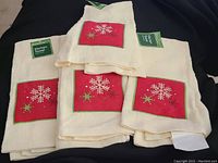 Five white kitchen towels with red square patches embroidered with white snowflakes and green holly berries, arranged on a dark fabric.