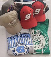 Several sports baseball caps and folded sports shirts including UNC Tar Heels and Boston Celtics.