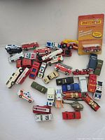 Top view of a collection of vintage toy emergency vehicles including fire trucks, ambulances, police cars, a helicopter, and other emergency vehicles assembled on a white surface.
