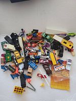 Overview photo showing variety of vintage toy vehicles and accessories scattered on surface including cars, trucks, construction vehicles, plastic figures, traffic cones and crate.
