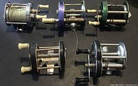 Top view of five antique fishing reels laid out side by side, showing different colors, handles, and fishing lines on one reel.