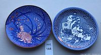 Two Asian decorative 12-inch porcelain plates side by side on a white surface. One is blue with pink chrysanthemums and cherry blossoms, the other blue and white with scaled background and detailed ship and gourd motifs.