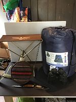 Camping survival lot displayed on black table including Fenwick folding stool, sleeping bag in blue sack, leather canteen with attached knife, and black binoculars.
