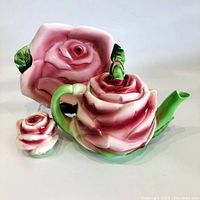 Front view of the rose-shaped ceramic teapot set with biscuit plate and shaker, highlighting the pink and green color scheme and detailed floral design.