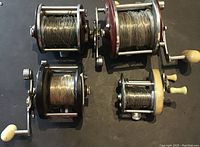 Four fishing reels laid out on dark surface, showing loaded steel fishing line on each spool. Three reels have Bakelite bodies with metal frames and cranks featuring cream-colored handles. One smaller reel is lighter with pale yellow handles.