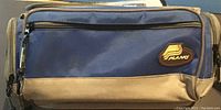 Close-up of the Plano fishing tackle bag showing blue and gray fabric with zippered compartments and Plano logo patch.
