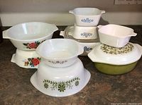 Overview of all milk glass baking casseroles showing various vintage floral patterns and sizes stacked or grouped on countertop.