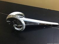 Full side view of the chrome vintage Dodge ram hood ornament placed on a black surface showing shape and wear.