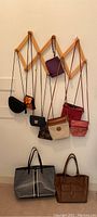 Photo showing 8 assorted women's handbags and clutches hanging on a wooden accordion rack against a white wall. Bags include neutral and bright colors, leather and fabric materials, and mix of sizes.