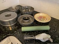 Bakeware and kitchen tools including pie pans, cake pans, rolling pin, and white hand mixer with beaters on black countertop
