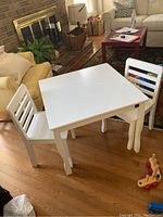 White children's square table with two matching chairs positioned around it in a room with hardwood floors and furniture.