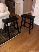 Two black wooden stools with rectangular seats on a wood floor and patterned rug