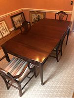 Photo showing full dining table with leaf extended and two chairs, set in a dining room with carpet and framed pictures nearby.