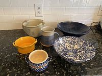 Seven ceramic items on kitchen counter including five bowls (yellow, blue floral, speckled, black, beige with blue floral) and two pitchers (gray/blue jug and beige bowl/pitcher)
