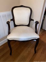 Full front view of antique armchair showing white striped upholstery and dark carved wood frame with armrests.