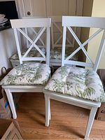 Front view of two white cross back dining chairs with green and white patterned cushions on wooden floor.