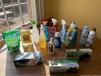 Photo of assorted cleaning liquids and sprays such as dish soap, glass cleaner, surface cleaner, and trash bags placed on a wood table