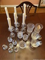 Collection of crystal candle holders with white taper candles, crystal salt and pepper shakers, small crystal bowls, glass bowls, and decorative scalloped vases shown on a wooden table.
