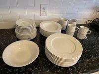 Full view of most of the dinnerware stacked on kitchen counter showing the set including plates, bowls, and cups with embossed leaf garland pattern.