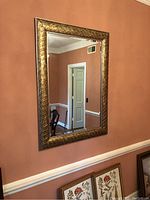 Large rectangular wall mirror with an ornate gold finish frame mounted on a peach-colored wall, showing part of the room and furniture reflected.