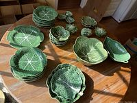 Multiple green glazed ceramic dishes in the shape of cabbage leaves, including various sizes of plates, bowls, and lidded bowls, arranged on wooden table.