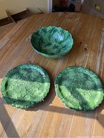 Photo showing the full set of a green leaf patterned ceramic serving bowl and two dinner plates on a wooden table, with natural light and some background elements.