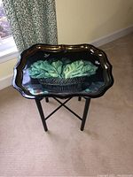 Full view of Ian Logan metal frame tray table on beige carpet, showing tray top with basket of leafy vegetables design and black folding legs.