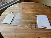 Three cutting boards: a small white plastic one with handle, a large transparent glass cutting board with rubber feet at corners, and a large white plastic cutting board with handle.