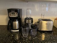 Photo showing an Oster 2-slice toaster, Ninja blender base with 3 blending cups and lids, and a Cuisinart coffee maker with thermal carafe placed on a black granite kitchen countertop against a white tiled backsplash.