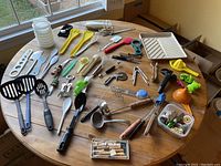 Assorted kitchen utensils and gadgets spread on a wooden table, showing variety of spatulas, peelers, ladles, tongs, and small kitchen tools mainly plastic and some metal.