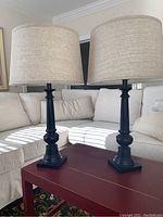 Pair of black lamps on a red table with beige couch background, showing full height and style.