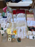 Full assortment of linens including napkins, runners, embroidered cloths, crystal coasters, and napkin rings arranged on a beige carpet.