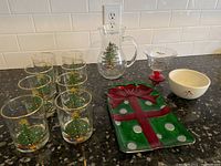 Eight Christmas tree decorated glasses, one Christmas tree pitcher, dessert glass, and white bowl, rectangular green tray with red bow pattern on black granite counter