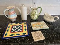 Kitchen counter view of 3 ceramic pitchers, 1 gravy boat, and 3 trivets arranged side by side.