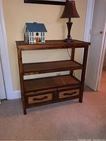 Front view of wooden shelf unit with wicker shelves and two drawers, showing its rustic style and placement next to a wall with a lamp and a decorative house on top.