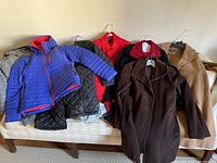 Six various women's winter coats arranged on a couch, showing different styles and colors including blue, red, black, brown, tan, and gray coats.