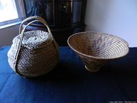 Photo showing two woven baskets on a dark cloth surface by a window. One round basket has a lid and handles; the other is a conical shaped basket with some purple dye markings.