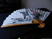 Side angle showing partially open fan with black and red floral painting on white canvas, wooden frame base visible.