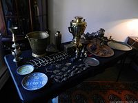 Overview of all assorted brass, copper and metal decorative items arranged on a black cloth-covered table, showing a variety of items including trays, samovar, candlesticks, bowls, hooks and figurines.