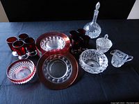 Photo showing assortment of clear and red-tinted cut glassware, including glasses, plates, decanter and sugar bowl on a dark cloth surface.