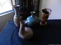 Overview of all four assorted large pottery vases placed on a black cloth with a window and cabinet in the background. Vases feature varied shapes and styles including multicolored bands, blue-green glaze, and handmade rustic forms.