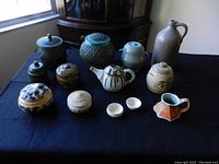 View of all 13 assorted handmade pottery items displayed on black cloth. Includes canisters, teapot, bowls, jug and various vessels with assorted glazes and textures.