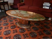 Oval coffee table with multicolor marble top and carved wood pedestal base on patterned carpet near red sofa.