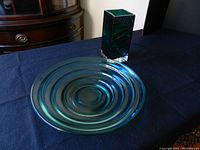 Green tinted circular glass platter with concentric ridges and dark green rectangular vase on black cloth surface.