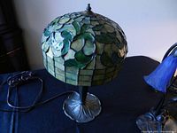 Full view of Tiffany style table lamp showing stained glass shade with green leaves and geometric squares; metal base with leaf design on a black cloth surface.