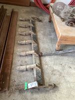 Full view of the metal front end loader teeth assembly laid on a concrete floor, showing eight teeth mounted on a horizontal metal bar with rust and wear.