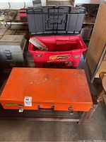 Photo showing three tool boxes stacked on a table: one large orange metal tool box in front, a red plastic toolbox behind it, and black plastic toolbox lid on top.