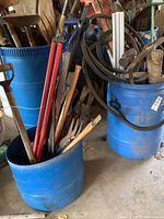 Two large blue barrels filled with various garden and building tools, mostly wooden and metal handles visible.