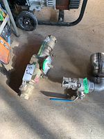 Photo showing both valves on the floor with a Watts brass valve featuring a pressure gauge and a stainless steel Pinacle valve with blue handle side-by-side.