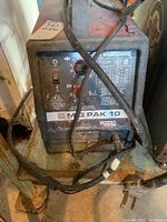 Front view of Lincoln Electric MIG PAK 10 with dial control, labels, and connected cables in dusty and used condition.